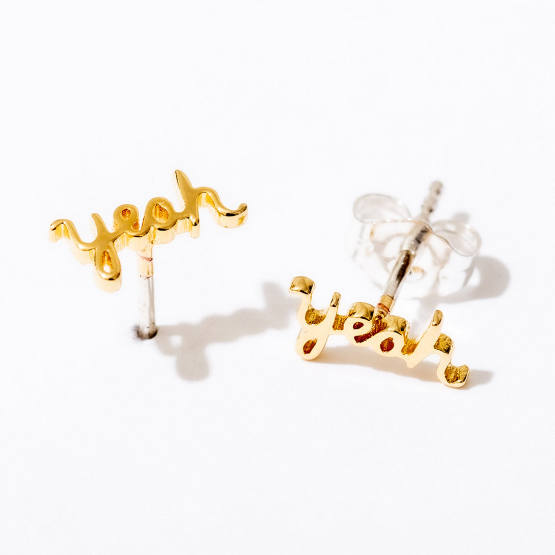 Gold Yeah Script Stud Earrings by Larissa Loden against a white background, casting a shadow