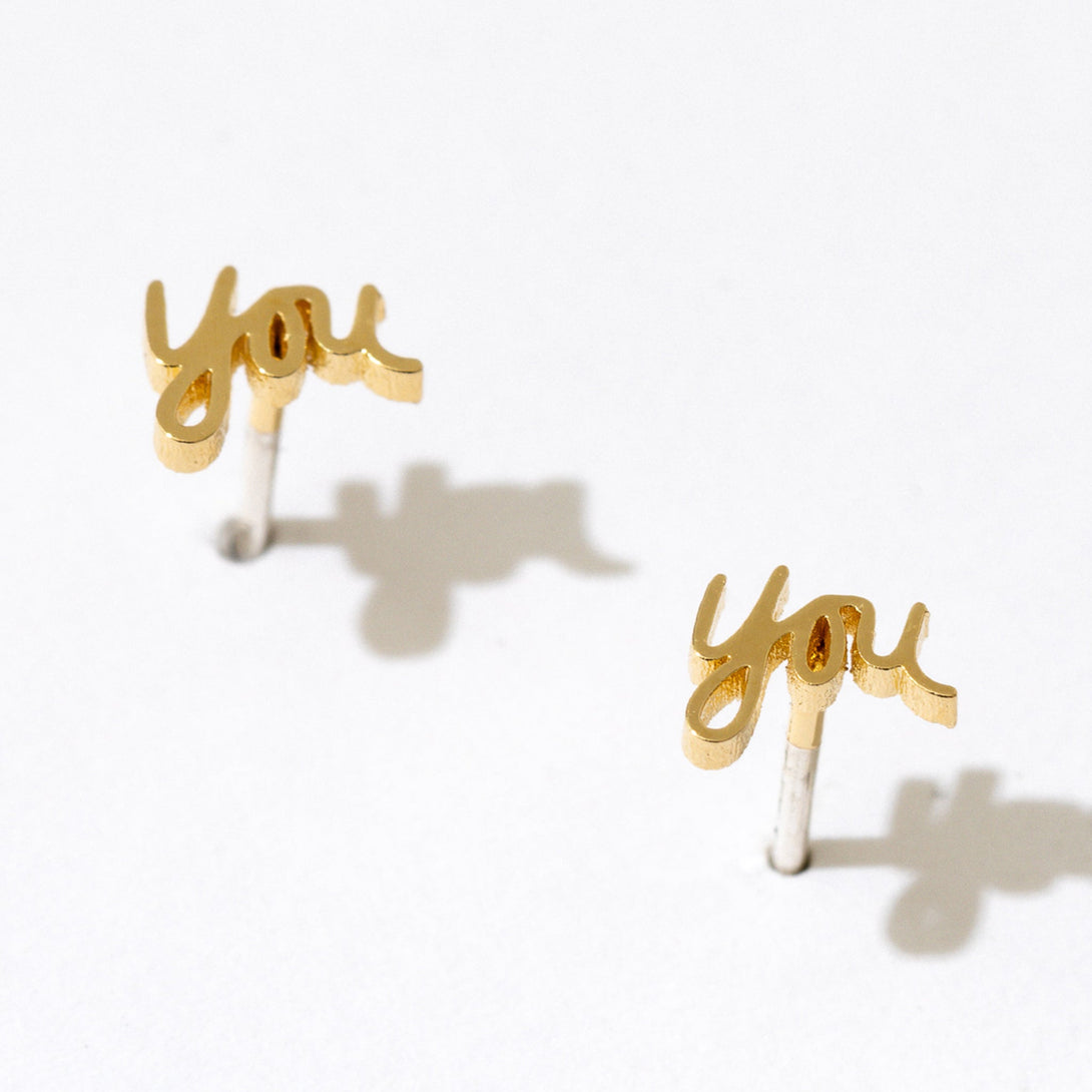 Gold You Script Stud Earrings by Larissa Loden against a white background, casting a shadow.