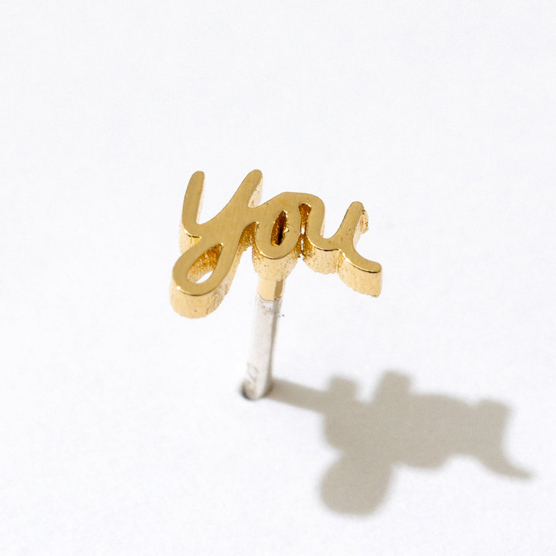 Single Gold You Script Stud Earring against a white background, casting a shadow